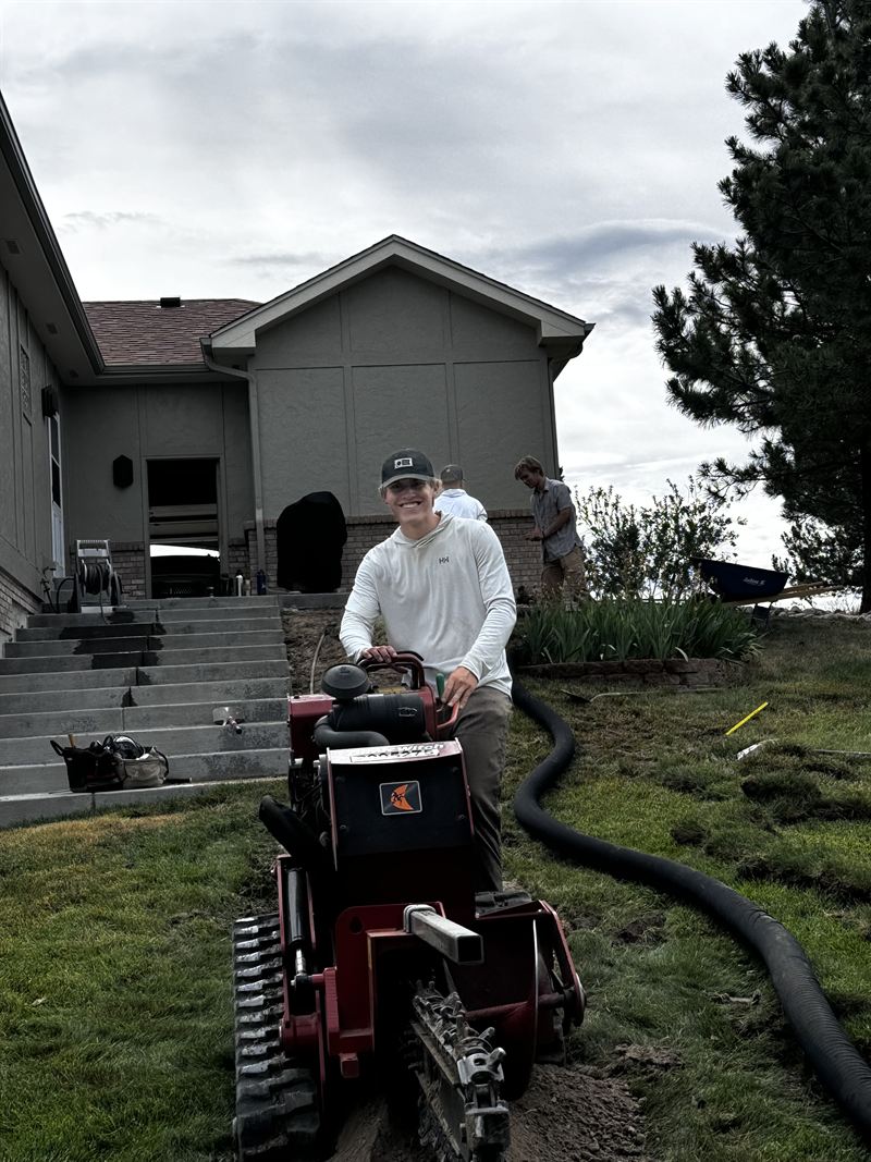 Mason smiling behind a walk-behind trencher on a lawn, with two crew members working by the house behind him
