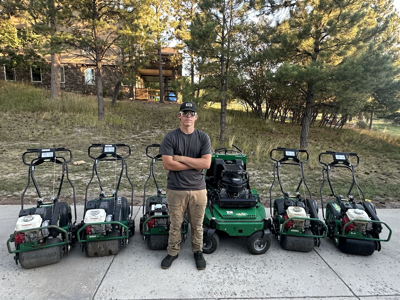 Mason standing on a sidewalk, arms crossed, with a lineup of five reel mowers and a stand-on mower
