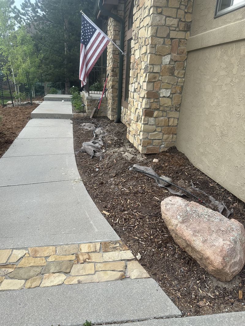 A front-walk bed stripped for new landscaping, with drip line exposed and a boulder in place